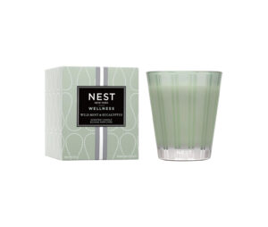 Green candle in a striped glass next to a patterned box labeled "NEST Wellness Wild Mint & Eucalyptus.