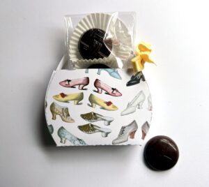 A decorative box featuring various shoe illustrations, with a chocolate treat and a yellow ribbon on top.