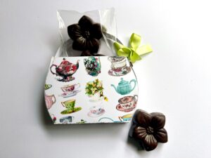 A decorative box featuring teacup illustrations, containing a chocolate flower and a clear bag of chocolates.