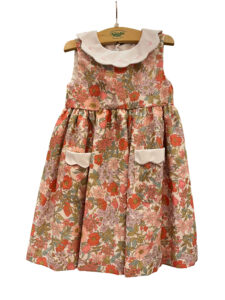 Floral dress in pink Liberty fabric with a white collar and two front pockets, hanging on a wooden hanger.