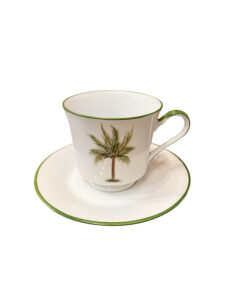 A white teacup with a palm tree design sits on a matching saucer.