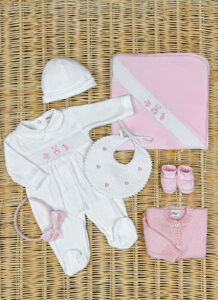 A white baby outfit, pink booties, a bib, a blanket, and a headband arranged on a woven surface.