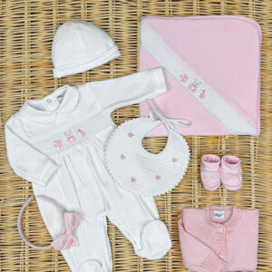 A white baby outfit, pink booties, a bib, a blanket, and a headband arranged on a woven surface.