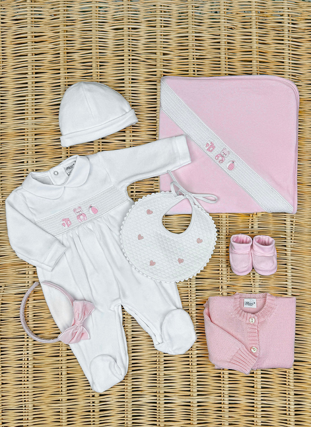 11489rosa_1_306b2951-8a45-4b30-972f-3b1af8d42dd5_1800x1800 A white baby outfit, pink booties, a bib, a blanket, and a headband arranged on a woven surface.