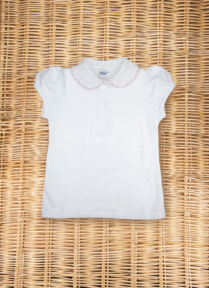 7957_ROSA_720x White short-sleeve polo shirt with a ruffled collar, laid flat on a woven surface.