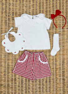 A white ruffled shirt, red gingham shorts, a bib, a headband, and a white sock arranged on a woven surface.