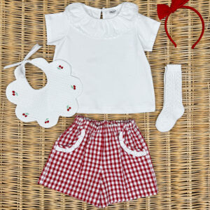 A white ruffled shirt, red gingham shorts, a bib, a headband, and a white sock arranged on a woven surface.