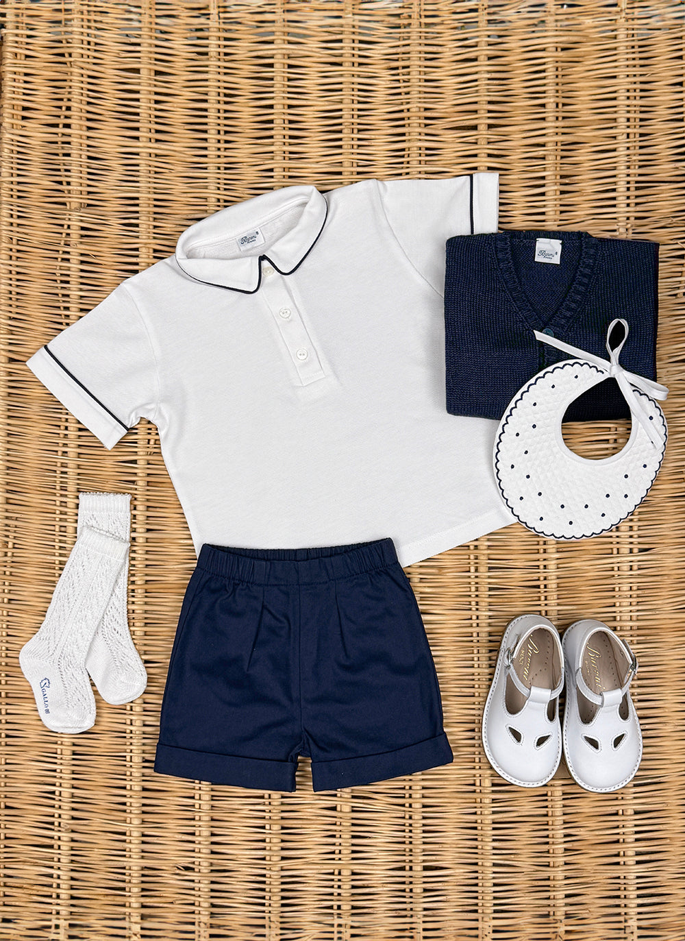 IMG_6530_1800x1800 A white polo shirt, navy shorts, a navy sweater, socks, a bib, and white shoes arranged on a woven surface.