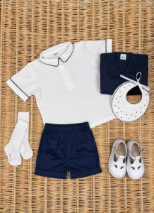 A white polo shirt, navy shorts, socks, a sweater, a bib, and white shoes arranged on a woven surface.