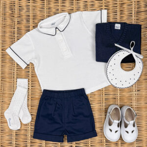 A white polo shirt, navy shorts, socks, a sweater, a bib, and white shoes arranged on a woven surface.