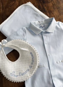 A light blue baby shirt, bib with embroidered text, and a matching blanket on a wooden surface.