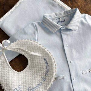 A light blue baby shirt, bib with embroidered text, and a matching blanket on a wooden surface.