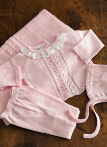 A pink knitted outfit with a ruffled collar and decorative patterns, laid out on a wooden surface.