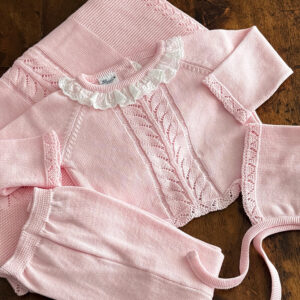 A pink knitted outfit with a ruffled collar and decorative patterns, laid out on a wooden surface.