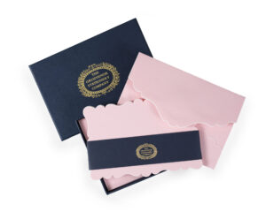 A navy box contains pink stationery with decorative edges and a gold logo band.