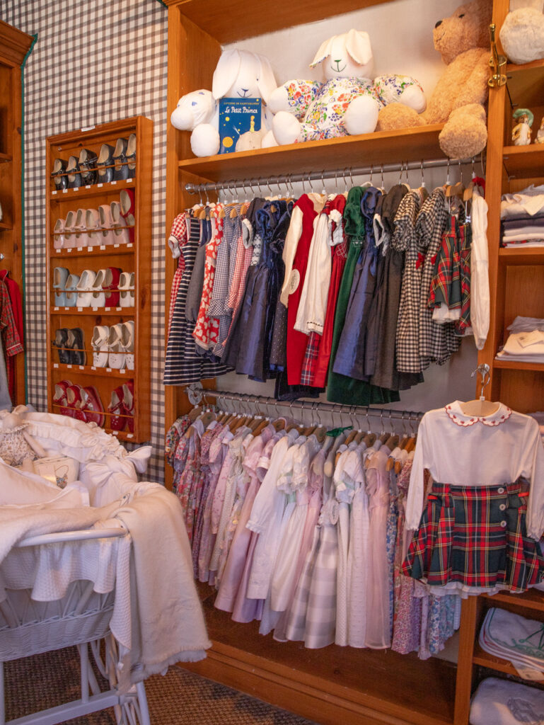 A wooden closet displays various children's dresses and shoes, with stuffed animals on top and a bassinet in front.