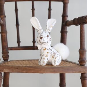 A decorative fabric bunny with a fluffy tail sits on a wooden chair.