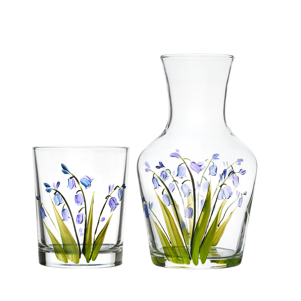 A glass carafe and matching tumbler, both decorated with bluebell flowers and green grass.