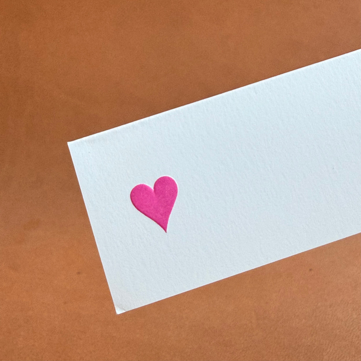 A white card with a pink heart shape on it, set against a brown background.