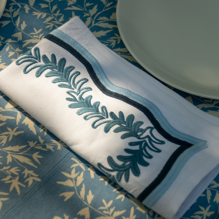 A folded napkin with blue embroidery rests on a patterned tablecloth, near a plate.
