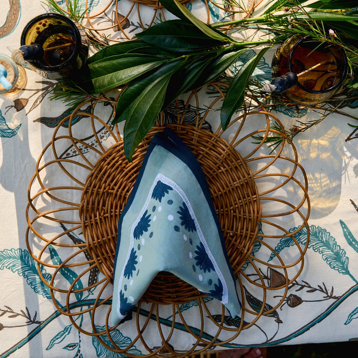A patterned napkin is folded on a woven placemat, surrounded by greenery and drinks.
