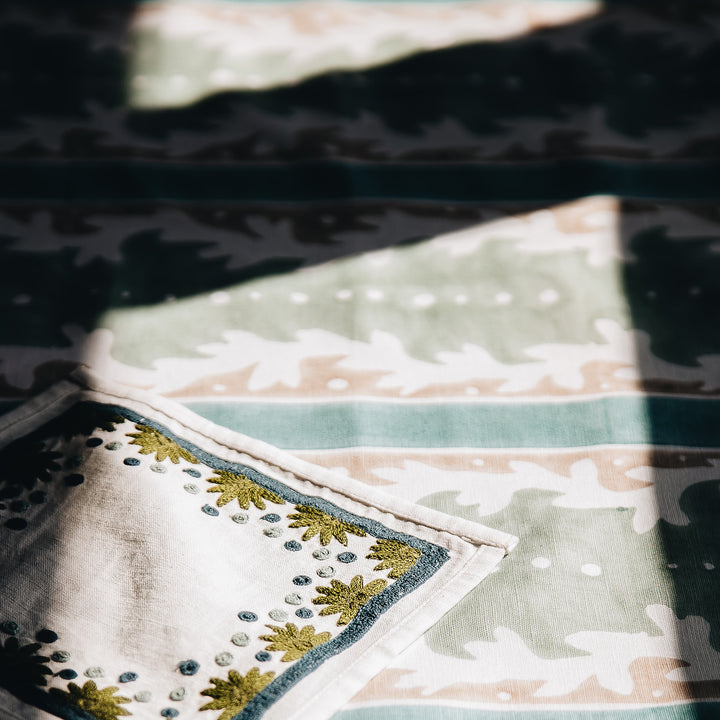 A decorative napkin with floral embroidery rests on a patterned tablecloth. Soft sunlight casts shadows.