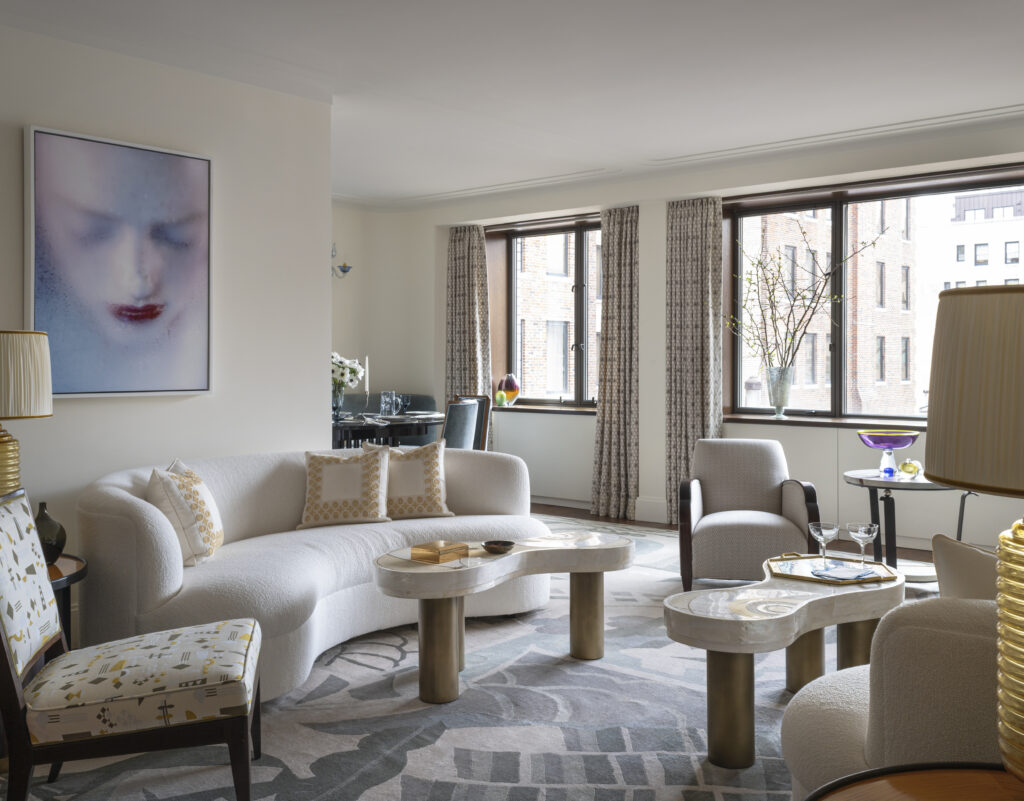 Bright living room featuring a curved white sofa, two armchairs, and a marble coffee table. Large windows with curtains.