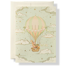 A whimsical hot air balloon floats among clouds, carrying a baby and colorful decorations.