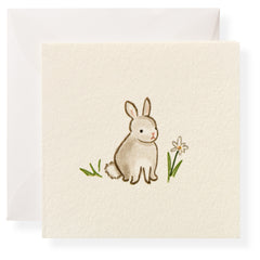 A cute brown rabbit sits beside a small flower and green grass on a textured card.