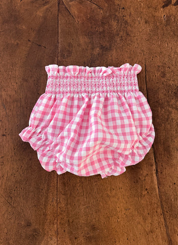 Pink gingham ruffled baby bloomers on a wooden surface.