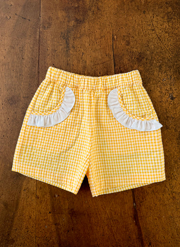 Yellow gingham shorts with white ruffled pockets on a wooden surface.