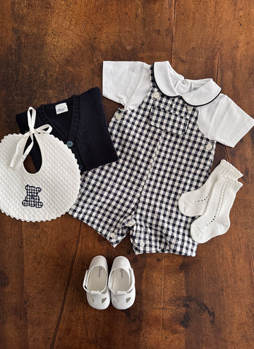 A baby outfit laid out: a navy cardigan, white shirt, checkered overalls, bib, socks, and shoes.