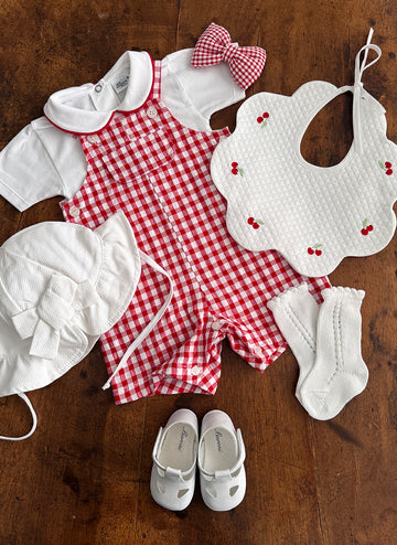 Red gingham romper, white hat, cherry-patterned bib, socks, and shoes arranged on a wooden surface.