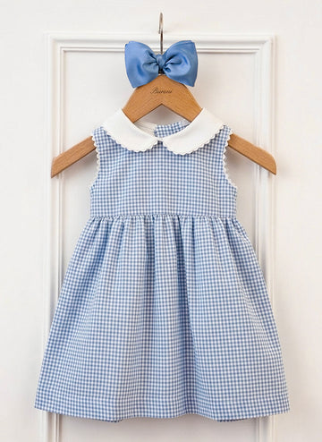A blue gingham dress with a white collar hangs on a wooden hanger, accompanied by a matching blue bow.