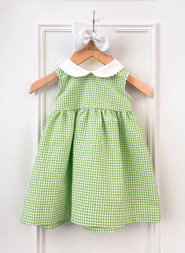 Green gingham dress with a white collar, hanging on a wooden hanger, accompanied by a white bow.