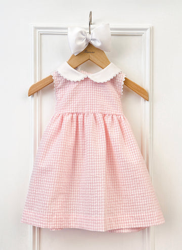 A pink gingham dress with a white collar hangs on a wooden hanger, topped with a white bow.